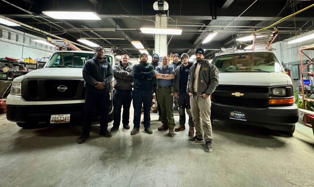 Seven workers stand in a garage beside two parked vehicles, surrounded by tools and equipment.
