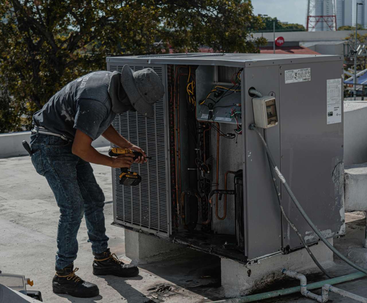 Technician repairs an air conditioning unit on a rooftop, using a power drill.