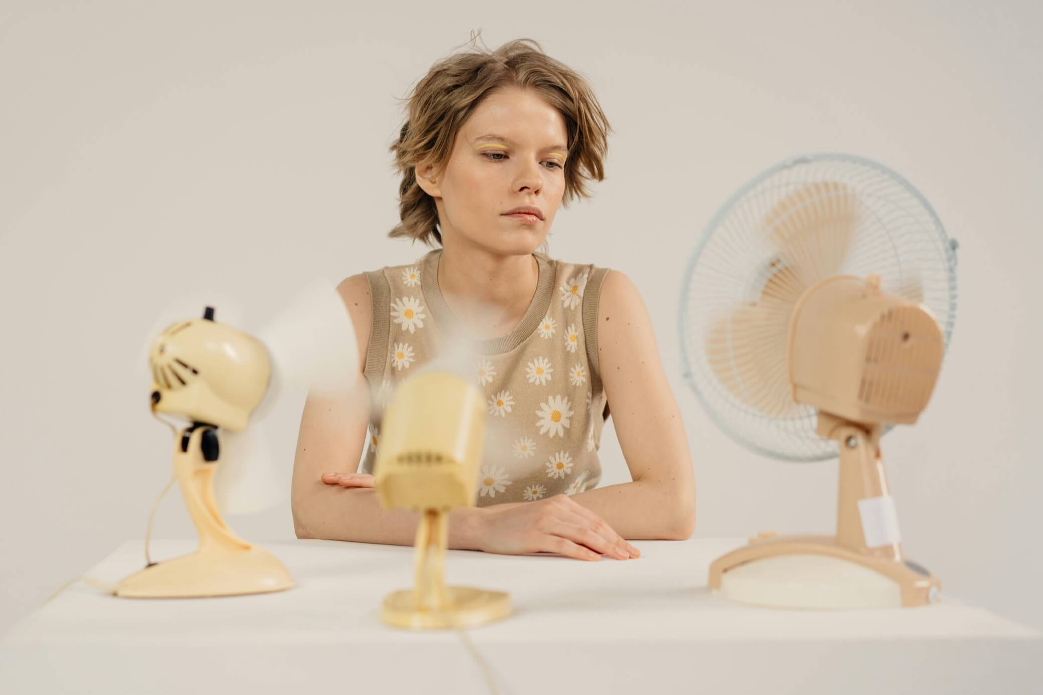 A woman sits calmly at a table, observing three stylish electric fans in pastel colors.