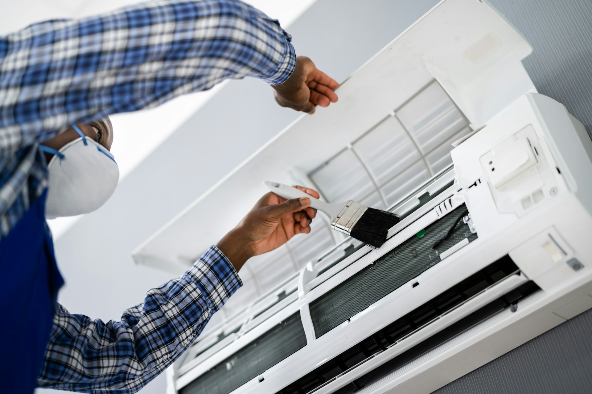 Man in a mask maintains an air conditioner using a paintbrush and wearing a blue plaid shirt.