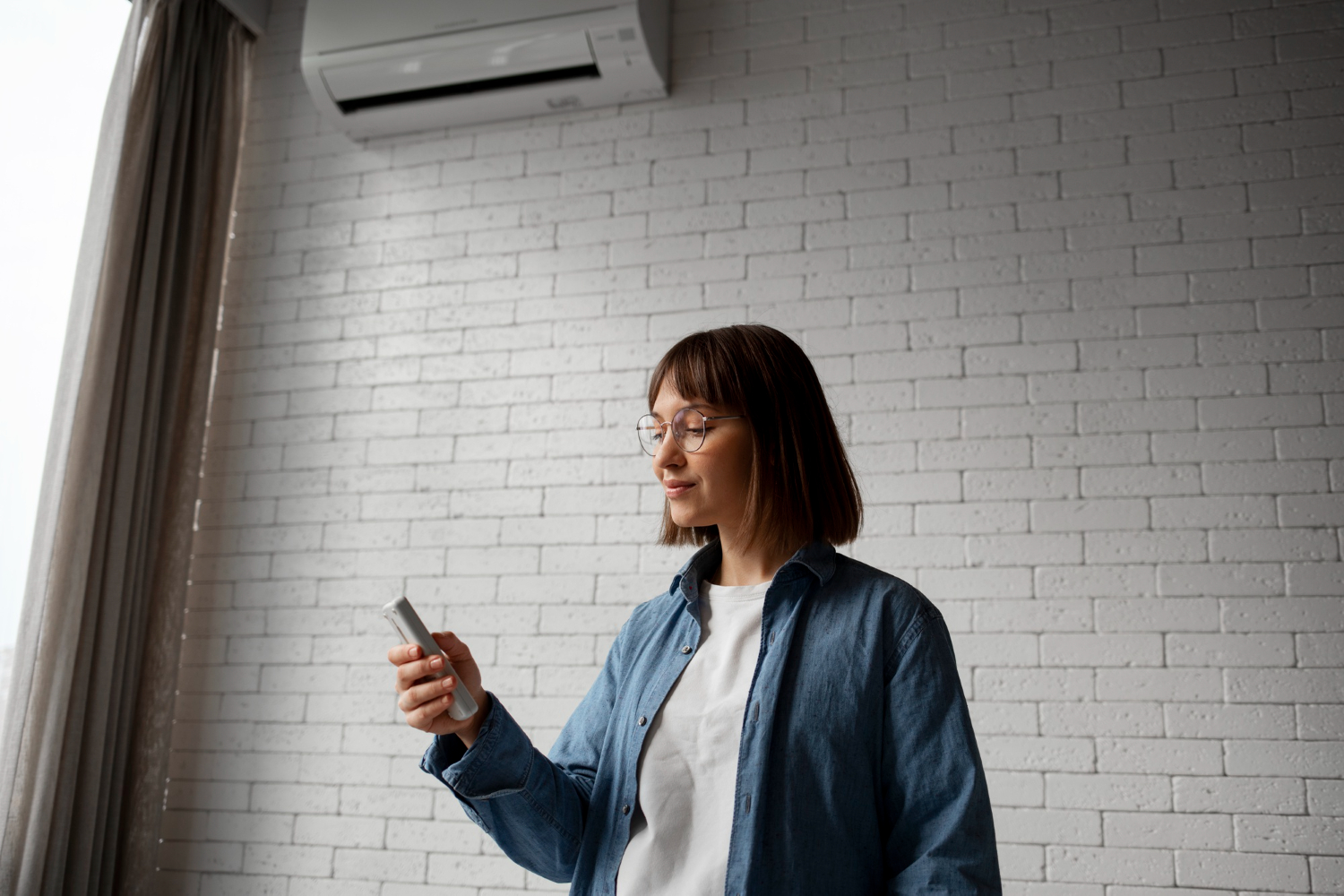 Young woman with glasses using a smartphone while standing beside a white brick wall.