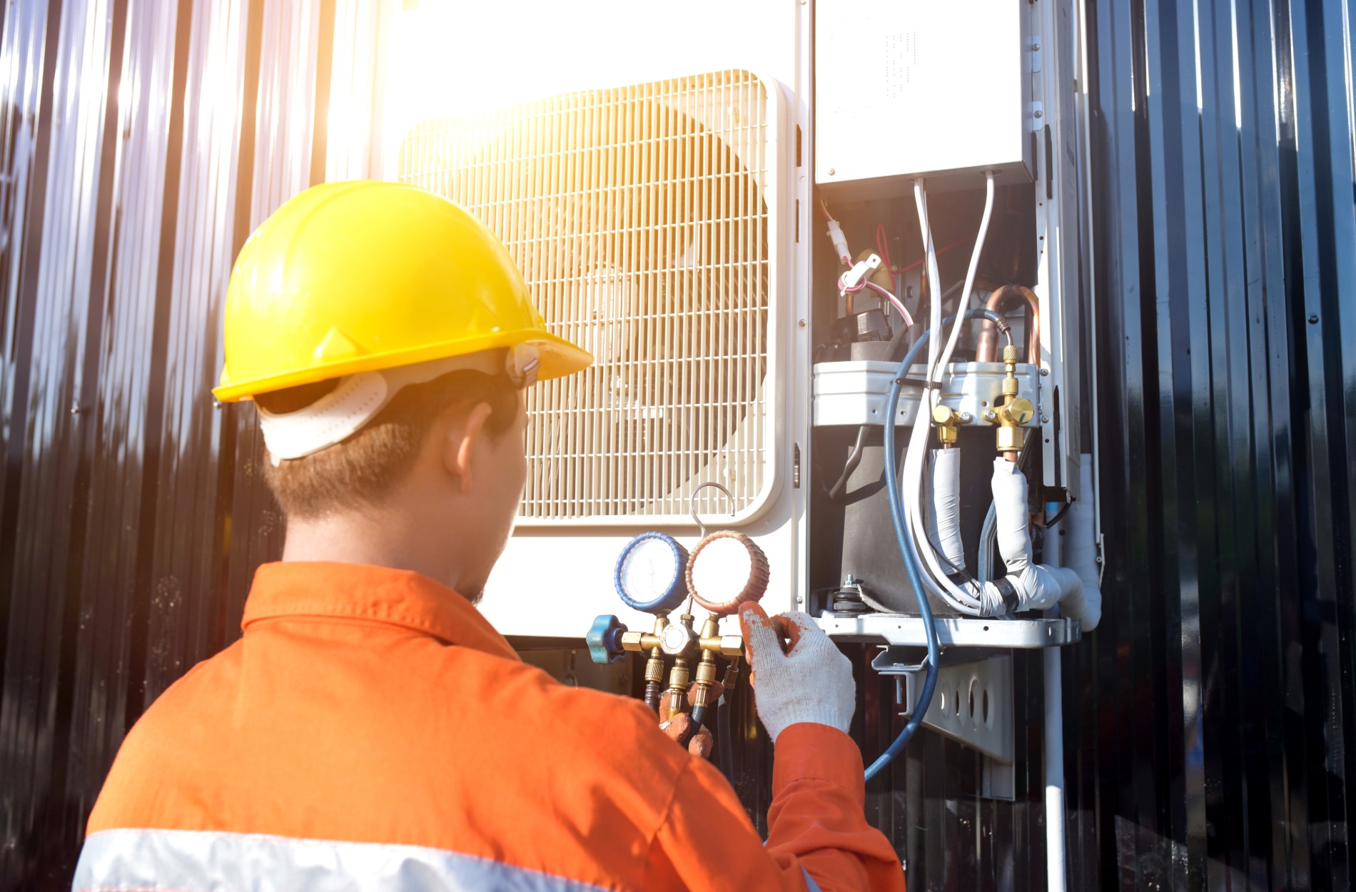 Technician in an orange uniform and yellow helmet adjusts HVAC gauges outdoors.
