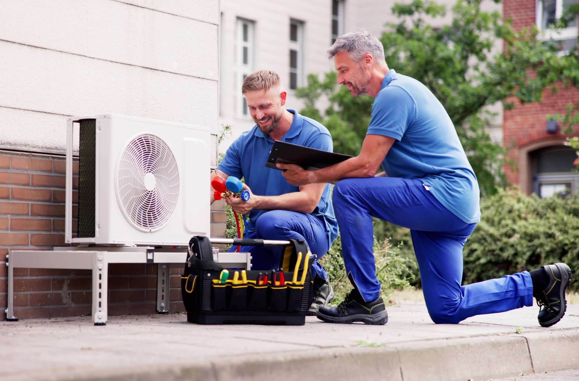 Two technicians in blue uniforms work on an outdoor air conditioning unit with tools.