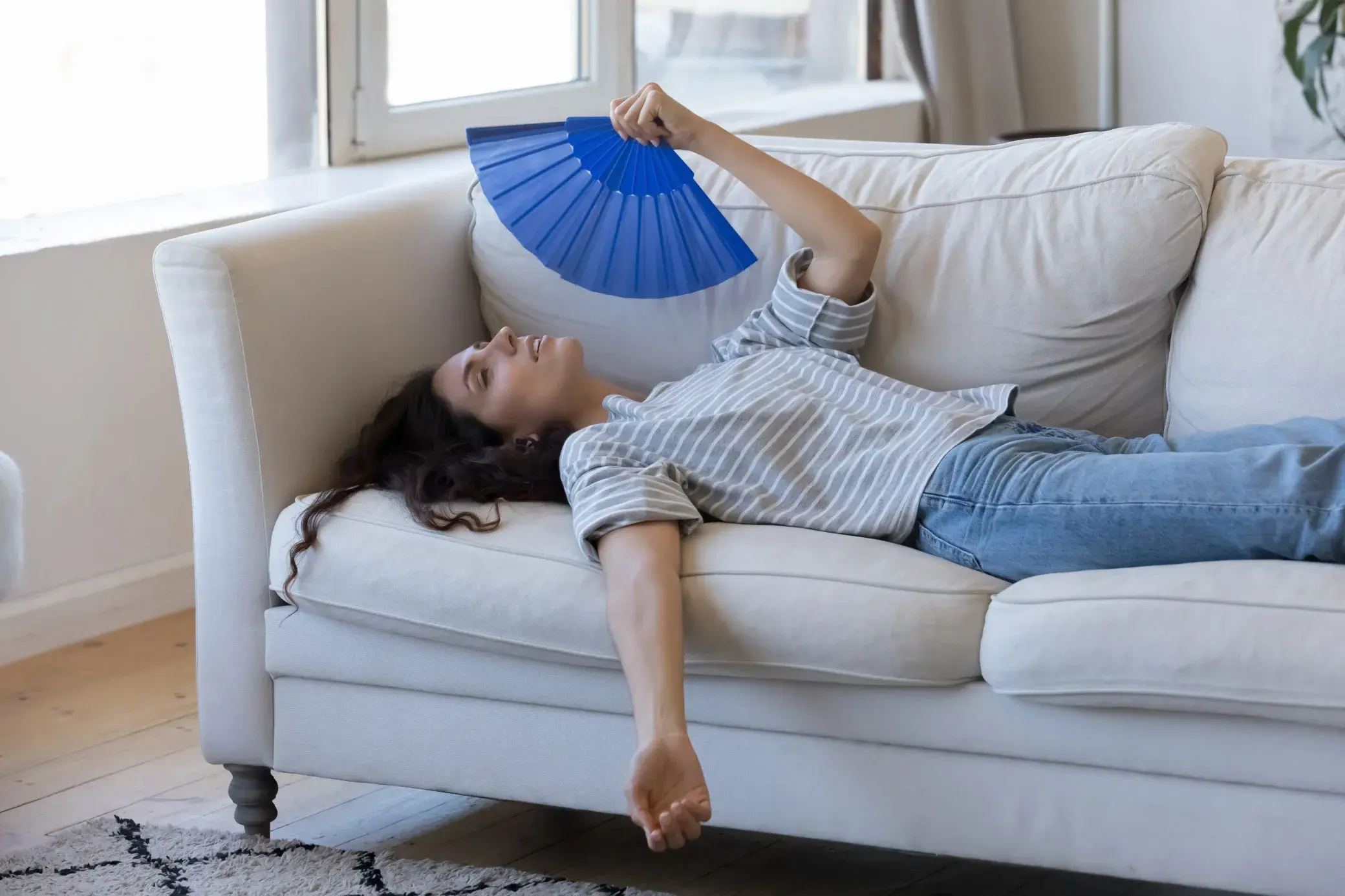 A woman lies on a sofa, holding a blue fan, relaxing in a bright, airy room.